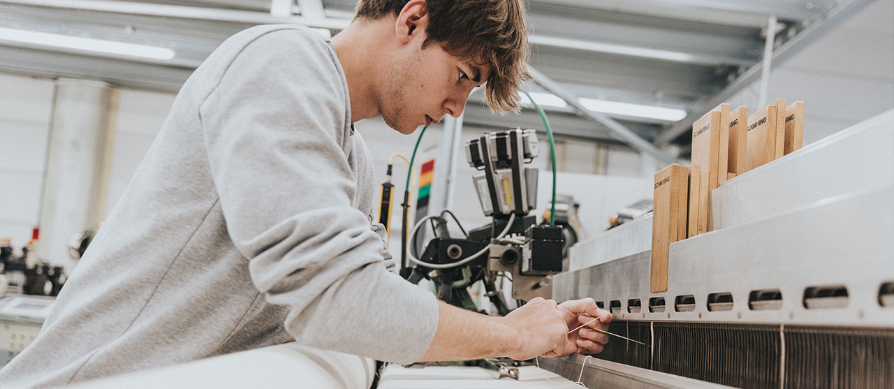 Student textieltechnologie van HOGENT haalt met een doorhaalhaak een kettingdraad door een hevel van een weefmachine met schachten.